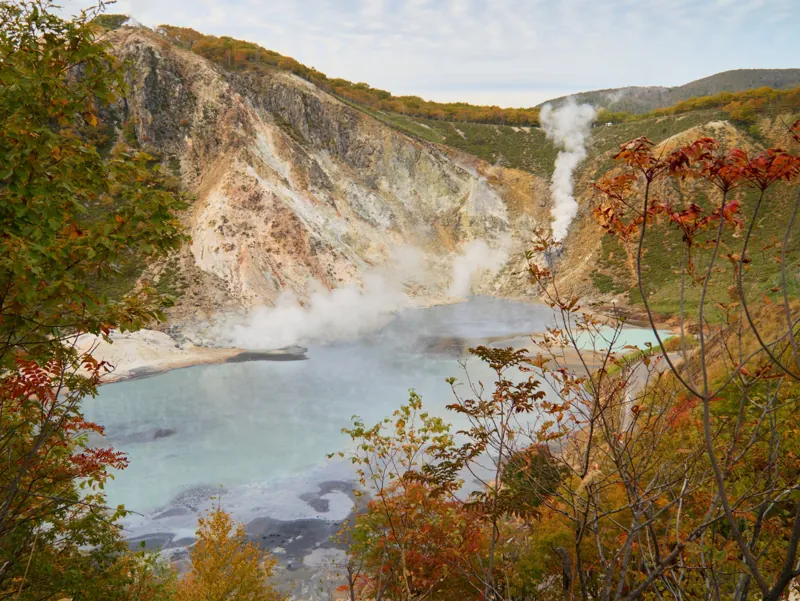 Un lago termale fumante in un cratere, tra le foglie d’autunno.