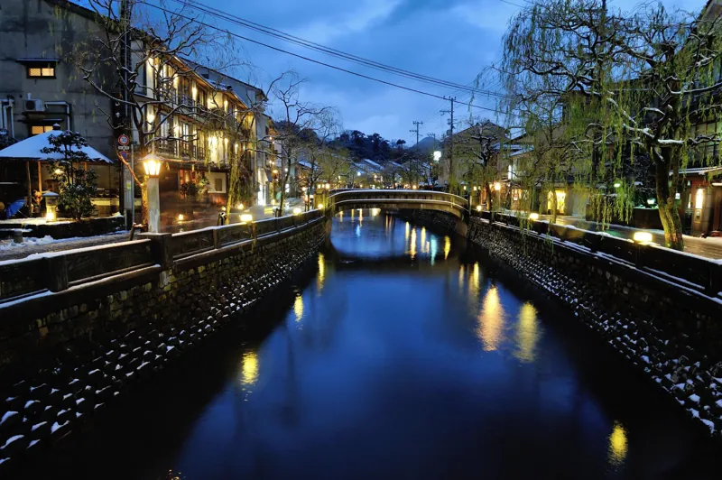 Il canale di Kinosaki sotto la neve all’ora blu, con i lampioni accesi e il ponte di pietra.